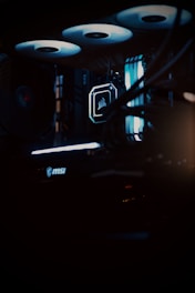 A close-up view of a computer gaming rig illuminated with blue LED lights. The interior shows components like cooling fans at the top, a glowing logo on a central component, and an MSI branded part at the bottom. The scene is dark, emphasizing the glow of the lights.
