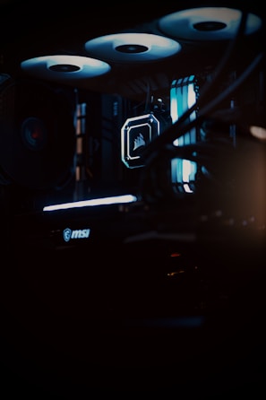 A close-up view of a computer gaming rig illuminated with blue LED lights. The interior shows components like cooling fans at the top, a glowing logo on a central component, and an MSI branded part at the bottom. The scene is dark, emphasizing the glow of the lights.