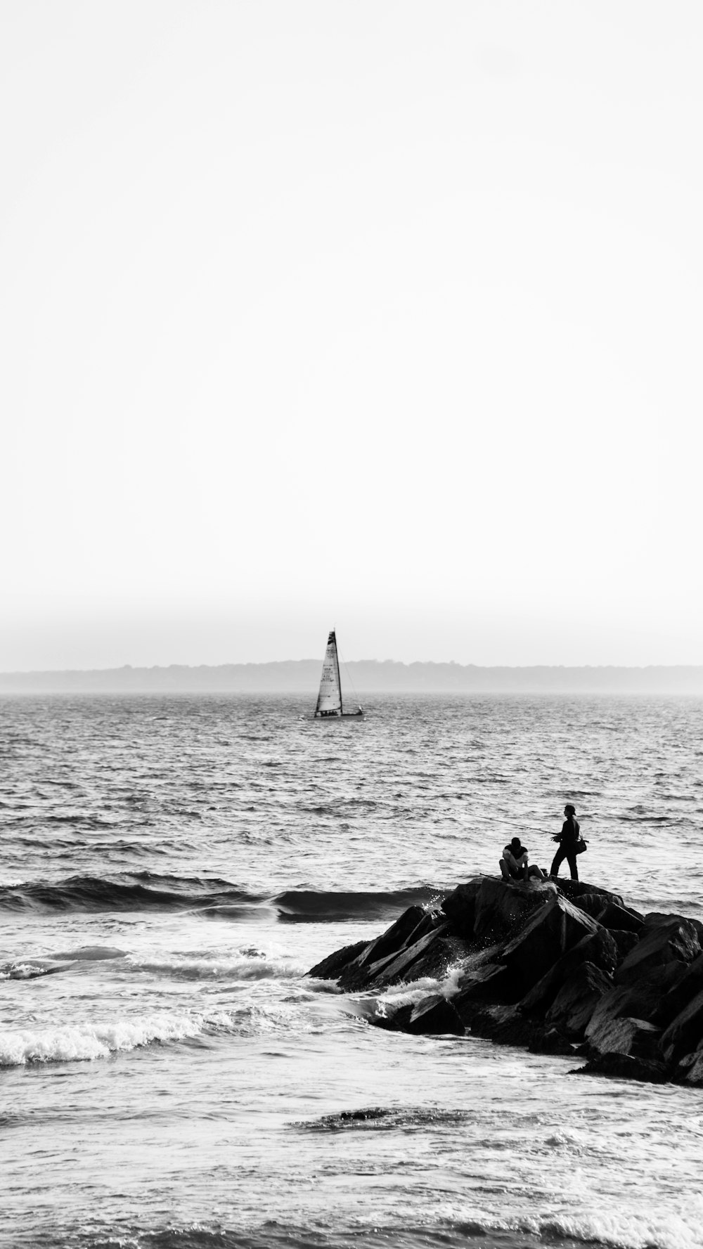 a man standing on a rock in the ocean with a sailboat