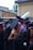 A group of graduates dressed in black gowns with purple and blue accents are gathered together outdoors. They are wearing academic caps. One of them is holding a metallic balloon. The background appears to be an urban setting with buildings and some greenery.