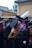 A group of graduates dressed in black gowns with purple and blue accents are gathered together outdoors. They are wearing academic caps. One of them is holding a metallic balloon. The background appears to be an urban setting with buildings and some greenery.
