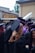 A group of graduates dressed in black gowns with purple and blue accents are gathered together outdoors. They are wearing academic caps. One of them is holding a metallic balloon. The background appears to be an urban setting with buildings and some greenery.