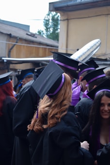 A group of graduates dressed in black gowns with purple and blue accents are gathered together outdoors. They are wearing academic caps. One of them is holding a metallic balloon. The background appears to be an urban setting with buildings and some greenery.