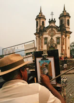 An artist, wearing a straw hat, is painting a detailed depiction of a historic church with twin towers and a cross on top. The painting is set up on an easel, and the artist is focused on capturing the intricate architectural details. Cobblestone streets and stone walls surround the scene. Additional people are visible in the background, contributing to a bustling yet peaceful atmosphere.