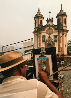 An artist, wearing a straw hat, is painting a detailed depiction of a historic church with twin towers and a cross on top. The painting is set up on an easel, and the artist is focused on capturing the intricate architectural details. Cobblestone streets and stone walls surround the scene. Additional people are visible in the background, contributing to a bustling yet peaceful atmosphere.