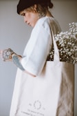 A person with tattoos wearing a black beanie and white shirt, holding a beige tote bag filled with white flowers. The tote bag has text printed on it, and the individual has several visible tattoos on their hand and neck.