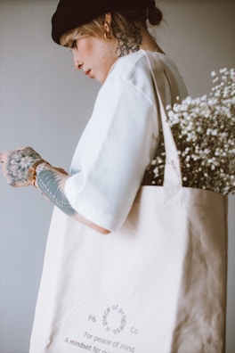 A person with tattoos wearing a black beanie and white shirt, holding a beige tote bag filled with white flowers. The tote bag has text printed on it, and the individual has several visible tattoos on their hand and neck.