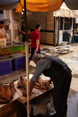 A bustling market scene with a vendor arranging fish on a wooden table. Several fish are displayed in neat rows, while other market goers, including a man in a red shirt, walk by in the background. The setting is under a canopy, providing shade to the market, with brick and tile walls visible.