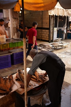 A bustling market scene with a vendor arranging fish on a wooden table. Several fish are displayed in neat rows, while other market goers, including a man in a red shirt, walk by in the background. The setting is under a canopy, providing shade to the market, with brick and tile walls visible.