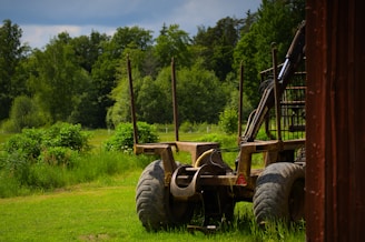 A rustic farm vehicle stands on a grassy field surrounded by lush green trees. The vehicle has large wheels and appears to be an agricultural implement or trailer situated next to a wooden structure on the right side.