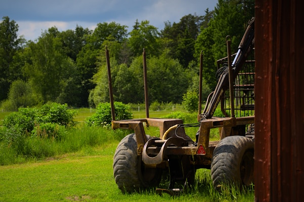 A rustic farm vehicle stands on a grassy field surrounded by lush green trees. The vehicle has large wheels and appears to be an agricultural implement or trailer situated next to a wooden structure on the right side.