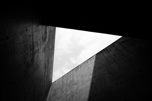 The perspective view of a concrete room with high walls forms a triangular aperture open to the sky. The surfaces of the walls are textured with a raw concrete aesthetic and are cast in varying shades of grey due to the play of light and shadow. The sky visible through the opening appears overcast, adding a sense of depth and contrast.