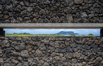 A stone wall with a narrow horizontal opening presents a view of a distant rocky island surrounded by vegetation and the sea. The stones vary in size and color, showcasing rugged textures and natural formations.