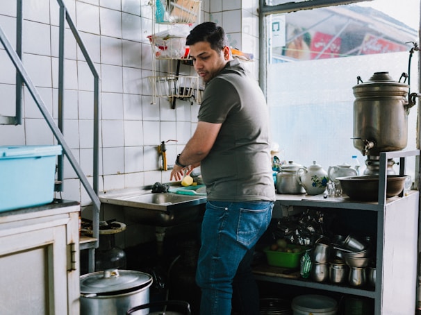 A person is washing vegetables at a kitchen sink, surrounded by various kitchen utensils and appliances. The room is quite compact, with tiled walls and shelves filled with pots, pans, and dishware. Natural light is coming through the window, adding to the overall brightness of the setting.