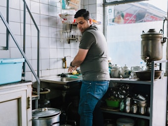 A person is washing vegetables at a kitchen sink, surrounded by various kitchen utensils and appliances. The room is quite compact, with tiled walls and shelves filled with pots, pans, and dishware. Natural light is coming through the window, adding to the overall brightness of the setting.