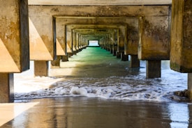 A symmetrical view under a concrete pier, showing a series of rectangular beams receding into the distance. The structure casts shadows on the water below. Waves gently lap against the sandy shore, with sunlight highlighting the green hues of the ocean.