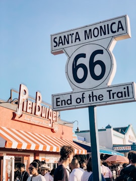 A large sign reading 'Santa Monica 66 End of the Trail' is prominently displayed in the foreground. Behind it is a restaurant named Pier Burger with a red and white striped awning. A crowd of people is gathered in front of the restaurant. The background shows additional buildings and storefronts, including one with 'Playland Arcade' signage.