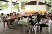 A group of people are sitting around two tables in a covered outdoor area, engaged in discussion. Some participants are wearing masks, and various electronic devices and papers are on the tables. The setting appears casual and informal.