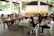 A group of people are sitting around two tables in a covered outdoor area, engaged in discussion. Some participants are wearing masks, and various electronic devices and papers are on the tables. The setting appears casual and informal.