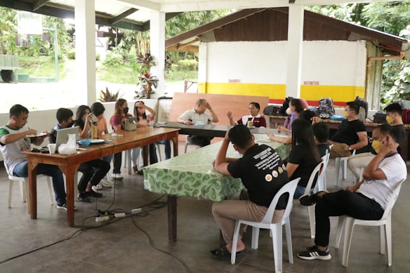 A group of people are sitting around two tables in a covered outdoor area, engaged in discussion. Some participants are wearing masks, and various electronic devices and papers are on the tables. The setting appears casual and informal.