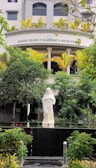 A serene statue of a religious figure stands amidst lush greenery in front of a building with the name Rajagiri College of Management and Applied Sciences. The statue is placed on a platform surrounded by well-maintained bushes and colorful flowers. The building features modern architecture with neutral colors and potted plants lining the balcony.