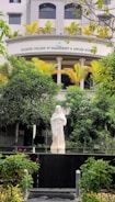 A serene statue of a religious figure stands amidst lush greenery in front of a building with the name Rajagiri College of Management and Applied Sciences. The statue is placed on a platform surrounded by well-maintained bushes and colorful flowers. The building features modern architecture with neutral colors and potted plants lining the balcony.