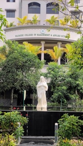 A serene statue of a religious figure stands amidst lush greenery in front of a building with the name Rajagiri College of Management and Applied Sciences. The statue is placed on a platform surrounded by well-maintained bushes and colorful flowers. The building features modern architecture with neutral colors and potted plants lining the balcony.