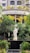 A serene statue of a religious figure stands amidst lush greenery in front of a building with the name Rajagiri College of Management and Applied Sciences. The statue is placed on a platform surrounded by well-maintained bushes and colorful flowers. The building features modern architecture with neutral colors and potted plants lining the balcony.
