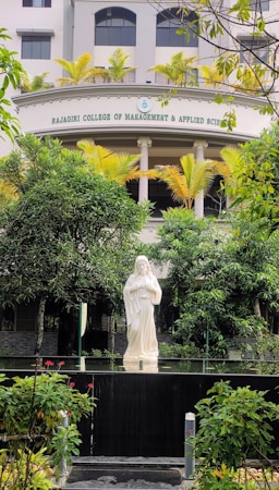 A serene statue of a religious figure stands amidst lush greenery in front of a building with the name Rajagiri College of Management and Applied Sciences. The statue is placed on a platform surrounded by well-maintained bushes and colorful flowers. The building features modern architecture with neutral colors and potted plants lining the balcony.