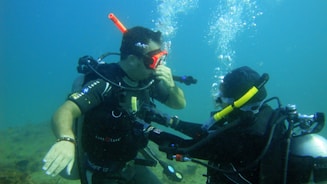 Two scuba divers are underwater, communicating with each other. They are wearing scuba gear, including masks, wetsuits, and oxygen tanks. Bubbles are rising above them as they exhale, and the water around them is clear with a slight blue tint.