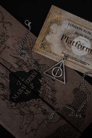 A collection of Harry Potter-themed items including a Marauder's Map and a triangular pendant with a circle inside, both resting on a dark surface. The map features intricate illustrations of buildings. A chain is visible around the pendant.