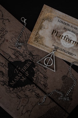 A collection of Harry Potter-themed items including a Marauder's Map and a triangular pendant with a circle inside, both resting on a dark surface. The map features intricate illustrations of buildings. A chain is visible around the pendant.
