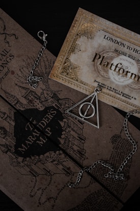 A collection of Harry Potter-themed items including a Marauder's Map and a triangular pendant with a circle inside, both resting on a dark surface. The map features intricate illustrations of buildings. A chain is visible around the pendant.