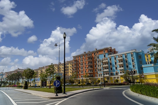 A vibrant street scene featuring colorful buildings with ornate balconies, surrounded by well-maintained greenery and palm trees. The sky is bright with fluffy clouds, creating a lively and cheerful atmosphere. Road signs and lampposts are visible along the curved road, which is smoothly paved.