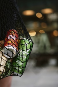 A close-up of a net bag containing a red can labeled 'Mate Mango' and a green can labeled 'Vegan Cola' with ingredients such as stevia. The background is blurred with warm bokeh lighting, contributing to a cozy atmosphere.