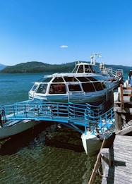 A sleek, modern boat docked at a wooden pier with a bright blue metal ramp leading to it. The surrounding area features a large, calm body of water and a backdrop of tree-covered hills. The sky is clear and bright, adding to the serene atmosphere. A couple of people are visible on the boat, appearing to be enjoying the view.