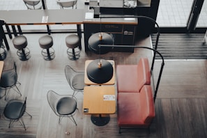 A modern cafe interior with a combination of wooden and metal furniture. The seating area includes metal wire chairs with black cushions and a small table with a red leather booth. Overhead lighting fixtures with black shades are hanging above the tables. The floor is covered with wood-like tile, and there are high stools lined up along a bar counter in the background.