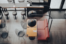 A modern cafe interior with a combination of wooden and metal furniture. The seating area includes metal wire chairs with black cushions and a small table with a red leather booth. Overhead lighting fixtures with black shades are hanging above the tables. The floor is covered with wood-like tile, and there are high stools lined up along a bar counter in the background.