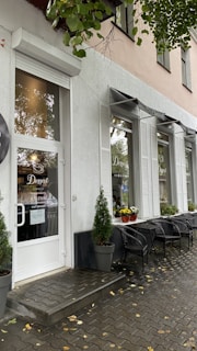 The exterior of a cafe features large windows with white shutters, a white door with signage, and potted plants beside the entrance. Outdoor seating includes several black chairs and tables on a paved walkway, with yellow flowers in planters along the windows.