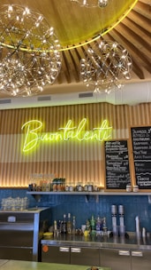A cozy café interior featuring a wooden ceiling with decorative hanging light fixtures. A neon sign reads 'Buontalenti' above a wooden counter with various bottles and items. A menu board in Russian lists different beverages.