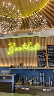 A cozy café interior featuring a wooden ceiling with decorative hanging light fixtures. A neon sign reads 'Buontalenti' above a wooden counter with various bottles and items. A menu board in Russian lists different beverages.