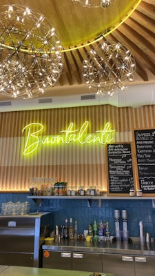 A cozy café interior featuring a wooden ceiling with decorative hanging light fixtures. A neon sign reads 'Buontalenti' above a wooden counter with various bottles and items. A menu board in Russian lists different beverages.