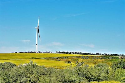 A calm rural landscape with a single wind turbine standing tall against a blue sky.