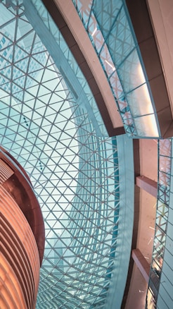 An intricate architectural interior featuring a geometric glass ceiling and a combination of curved and straight structures. The design is modern and expansive, showcasing the interplay of light and glass with metallic and wooden elements.