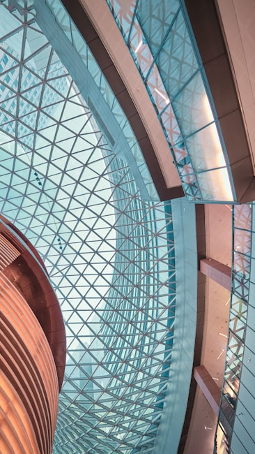 An intricate architectural interior featuring a geometric glass ceiling and a combination of curved and straight structures. The design is modern and expansive, showcasing the interplay of light and glass with metallic and wooden elements.