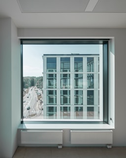 A modern office building with large windows is seen through a large glass window from inside another building. Behind the building, there is a road with cars and lush green trees in the background. The interior foreground includes a pair of white radiators beneath the window.
