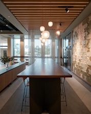 A modern interior space featuring a long wooden table with high chairs, hanging spherical pendant lights, and a geometric patterned wall. There are large windows allowing natural light to enter, and plants in vases on a side console. The ceiling has wooden slats and recessed lighting.