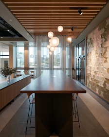A modern interior space featuring a long wooden table with high chairs, hanging spherical pendant lights, and a geometric patterned wall. There are large windows allowing natural light to enter, and plants in vases on a side console. The ceiling has wooden slats and recessed lighting.