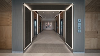 A modern hallway featuring two elevator doors on each side, with wooden paneling and sleek lighting accents. The floor is tiled, and directional signs are visible on the right side wall.