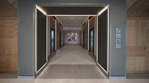 A modern hallway featuring two elevator doors on each side, with wooden paneling and sleek lighting accents. The floor is tiled, and directional signs are visible on the right side wall.